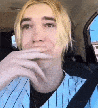 a young man with blonde hair is sitting in the back seat of a car with his hand on his mouth .