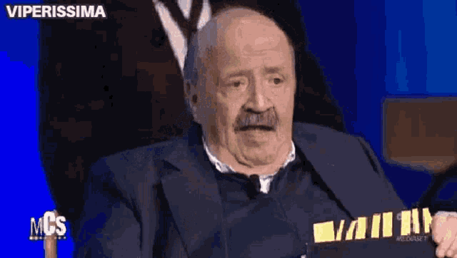 an elderly man with a mustache is sitting in front of a blue screen .