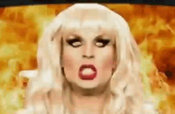 a drag queen with white hair and red lips is making a funny face in front of flames .