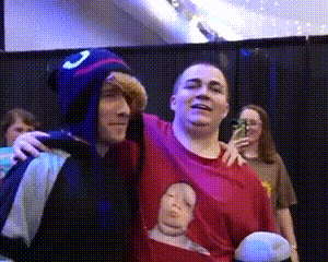 a man in a red shirt with a picture of a baby on it is hugging another man in a blue hat