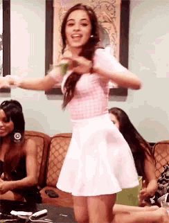 a woman in a pink top and white skirt is dancing in front of a group of women on a couch .