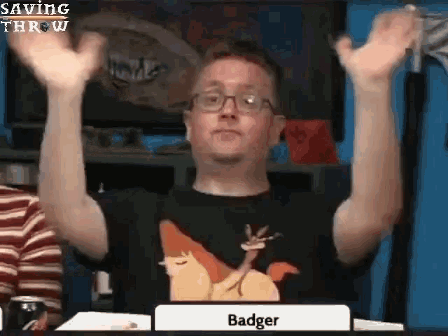 a man wearing glasses and a badger shirt is sitting at a table with his hands in the air .