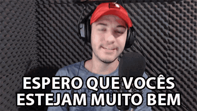 a man wearing headphones and a red hat says " espero que voces estejam muito bem " in front of a microphone