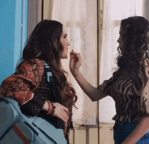 two women are standing next to each other in front of a window and one is touching the other 's face .