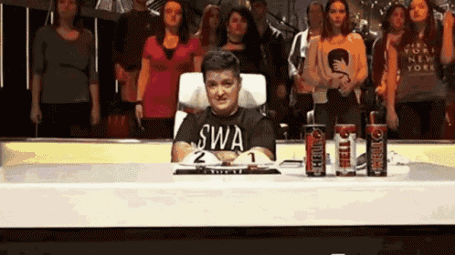 a man in a swa t-shirt sits at a desk in front of a crowd .