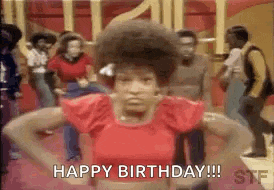 a woman in a red shirt is standing in front of a group of people dancing and saying `` happy birthday '' .