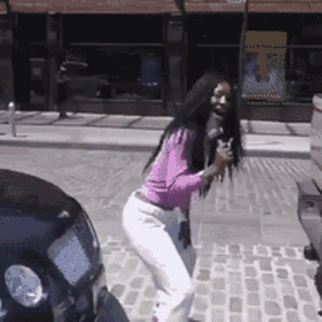 a woman in a pink shirt and white pants is standing on a sidewalk in front of a blue car