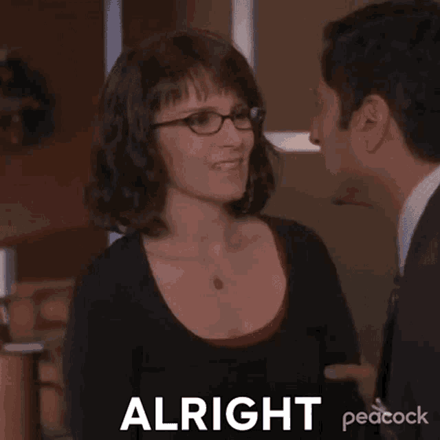 a woman wearing glasses is talking to a man in a suit and tie and says `` alright '' .