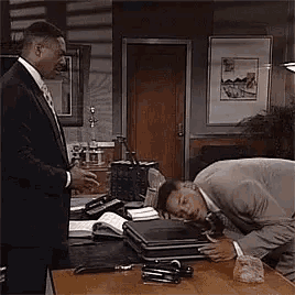 a man in a suit and tie is standing next to a man in a suit and tie laying on a desk .