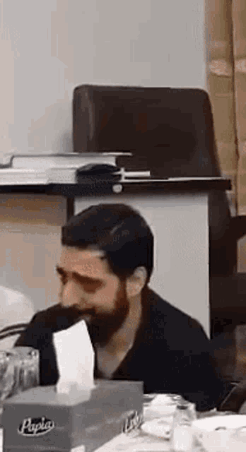 a man with a beard is sitting at a table with a tissue box .