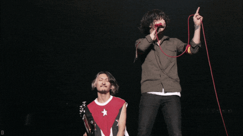 a man singing into a microphone while another man kneeling down looks on