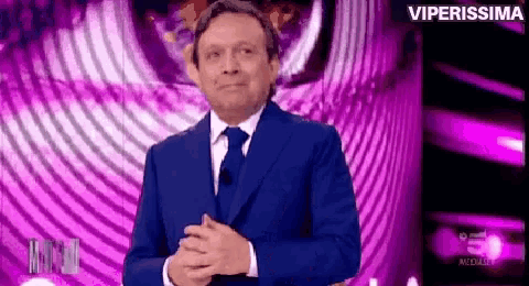 a man in a blue suit and tie is standing on a stage with his hands folded .