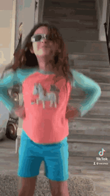 a little girl wearing sunglasses and a pink shirt is standing in front of a staircase .
