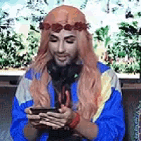 a man wearing headphones and a flower crown is sitting on a couch looking at his cell phone .