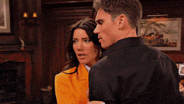 a man in a black shirt stands next to a woman in an orange jacket in front of a fireplace