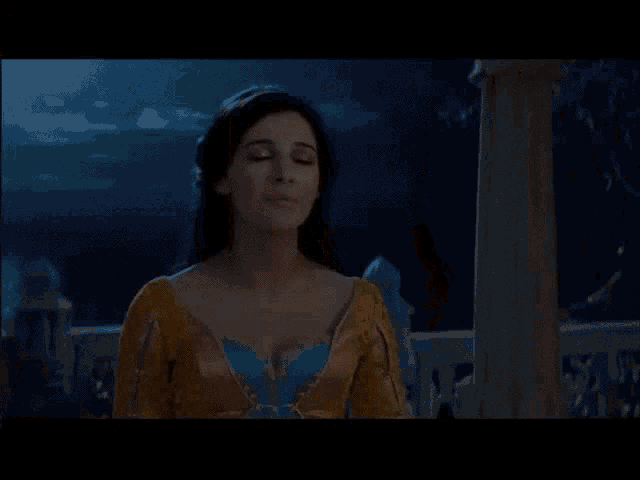 a woman in a yellow and blue dress stands on a balcony at night .