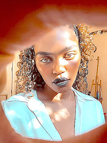 a woman with curly hair and black lipstick looks through a hole in a wall