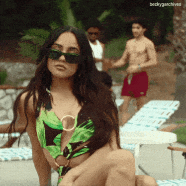 a woman in a green swimsuit sits on a chair by a pool with a man in red shorts behind her