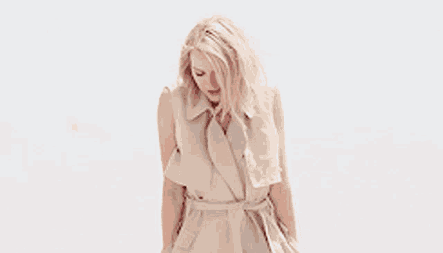 a woman wearing a tan trench coat is standing on a beach looking down .