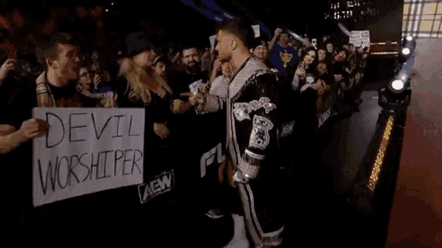 a man is standing in front of a crowd holding a sign that says `` devil worshiper '' .