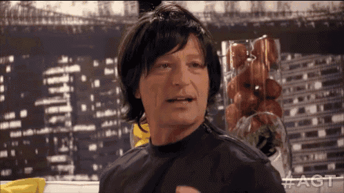 a man wearing a wig and a black cape is standing in front of a bowl of tomatoes .