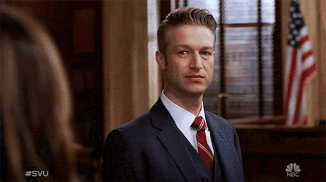 a man in a suit and tie stands in front of an american flag with the nbc logo on the bottom right