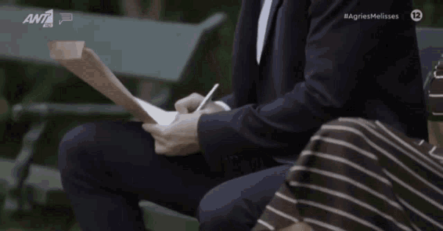 a man in a suit sits on a bench writing on a piece of paper in front of a sign that says agriesmelisses 12