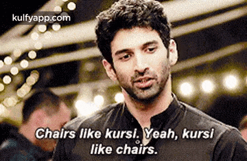 a man in a black shirt is talking about chairs and says `` chairs like kursi yeah , kursi like chairs . ''