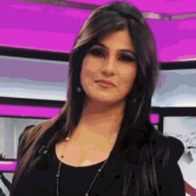 a woman with long dark hair is standing in front of a pink wall and looking at the camera .