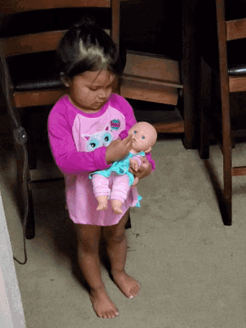 a little girl wearing a pink shirt with a cat on it holds a baby doll
