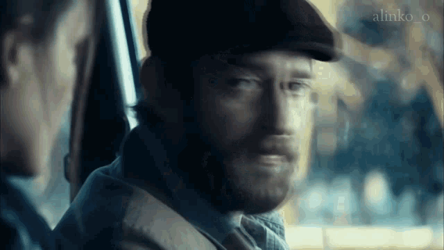 a man with a beard wearing a flat cap is sitting in a car with a woman