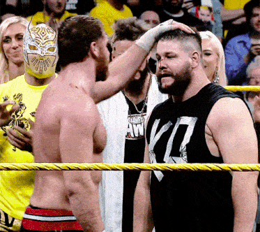 a wrestler wearing a black tank top with the letter k on it talks to another wrestler