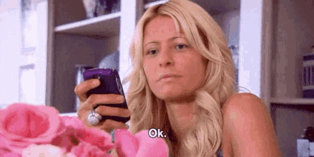 a blonde woman is holding a cell phone in her hand and saying ok .