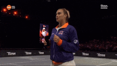 a woman stands on a tennis court wearing a blue and orange jacket