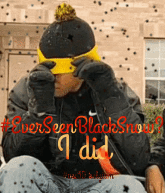 a man wearing a hat and gloves is sitting in front of a sign that says " ever seen black snow "