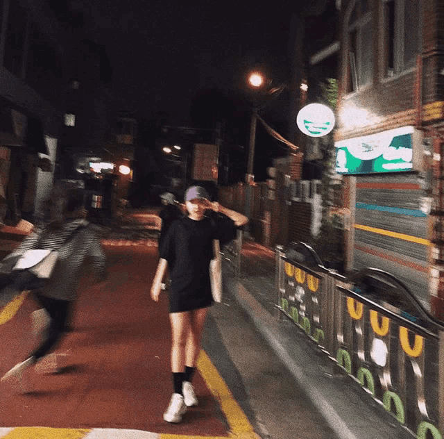 a woman walking down a street at night with a green sign that says ' a ' on it