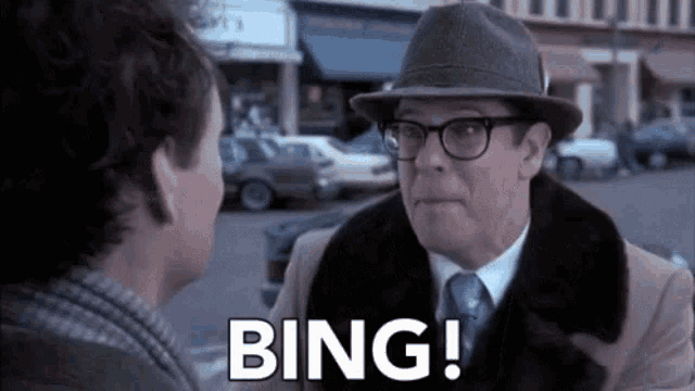 a man wearing a hat and glasses is talking to another man on a city street .