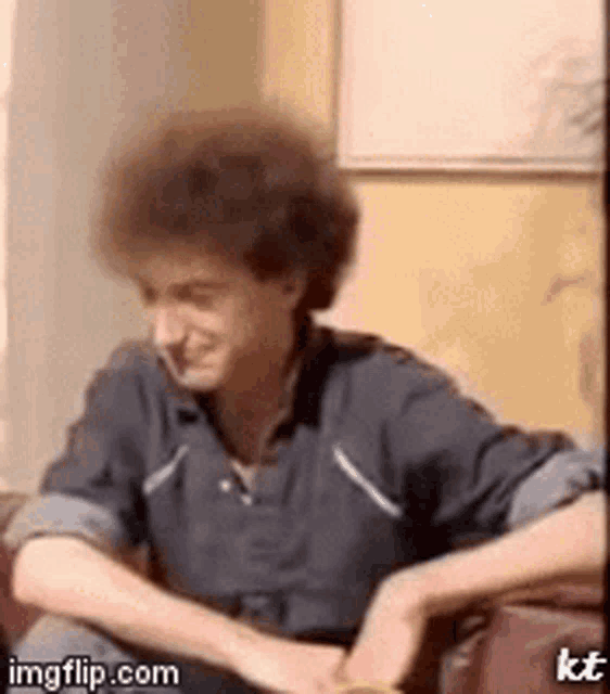 a man with curly hair is sitting on a couch with his hands folded .