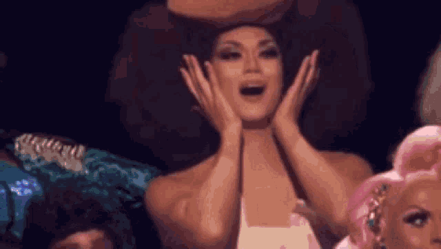 a drag queen with a large afro is holding her hands to her face and making a surprised face .