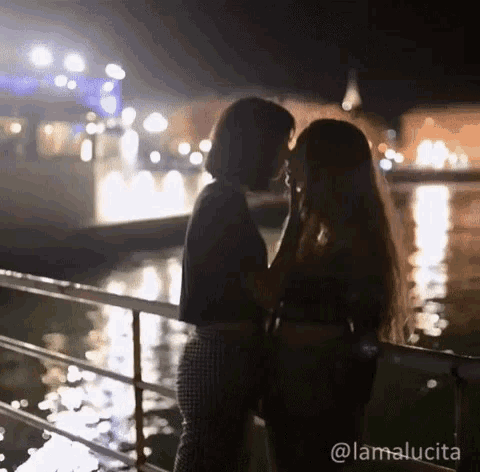 a couple of women are kissing in front of a body of water .
