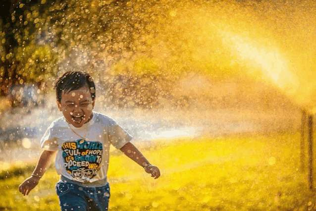 a young boy wearing a shirt that says " this world full of hope " is running through a sprinkler