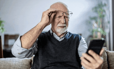 an elderly man wearing glasses is sitting on a couch looking at a cell phone .
