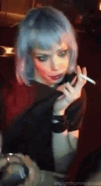a woman with blue hair is smoking a cigarette in a dark room .