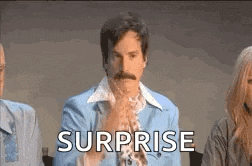 a man with a mustache is sitting in front of a group of people with the word surprise .