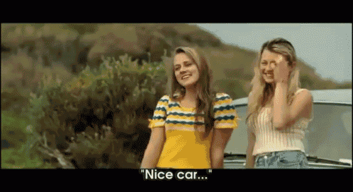 two women are standing next to each other in front of a car and one of them is saying nice car .