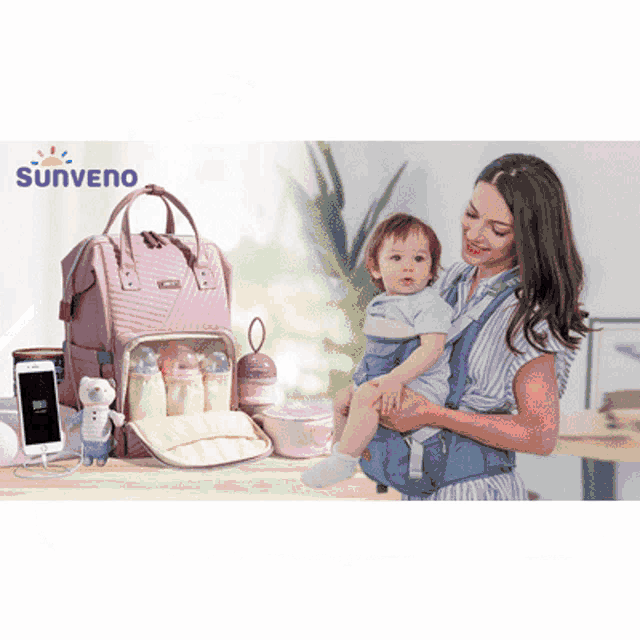 a woman is holding a baby in a carrier with a sunveno diaper bag behind her