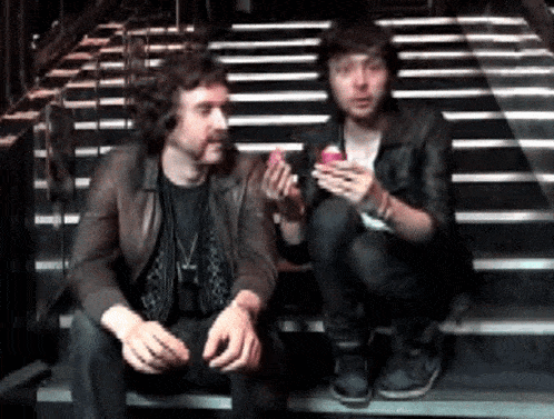 two men are sitting on a set of stairs one is eating a cupcake