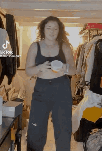 a woman in a black tank top and black pants is standing in a messy room holding a cup .
