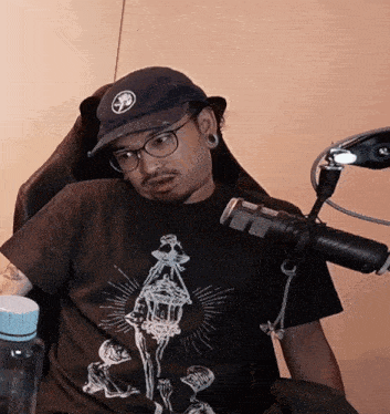 a man wearing a hat and glasses sits in front of a microphone and a water bottle