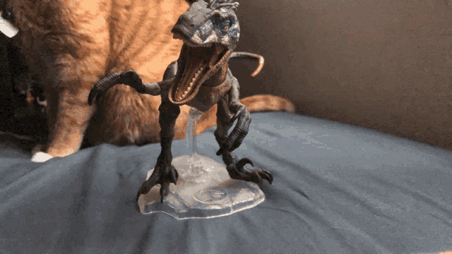 a toy dinosaur with its mouth open is on a bed next to an orange cat